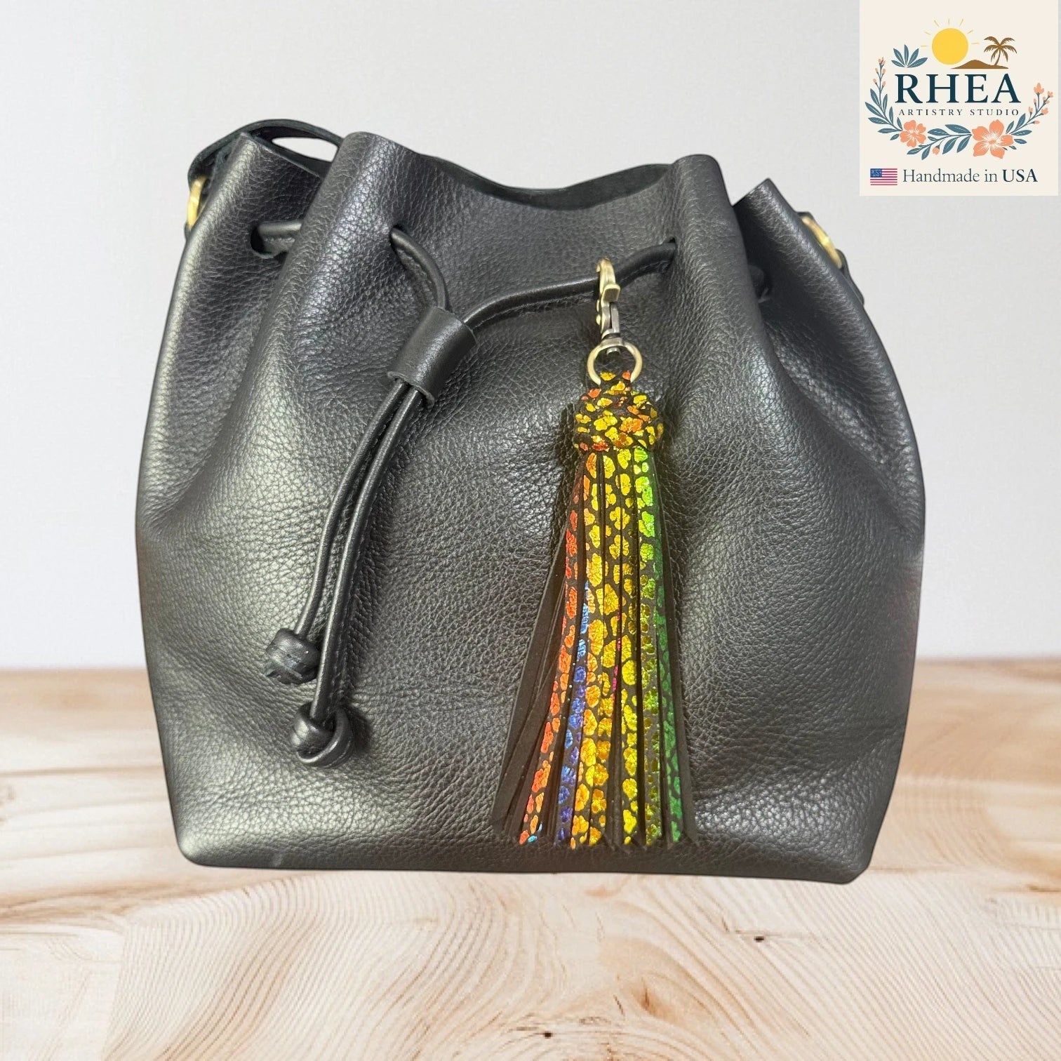 Rhea Artistry Studio handmade double braid leather tassel keychain with rainbow sparkle detail and brass lobster clasp attached to a black leather drawstring bag.