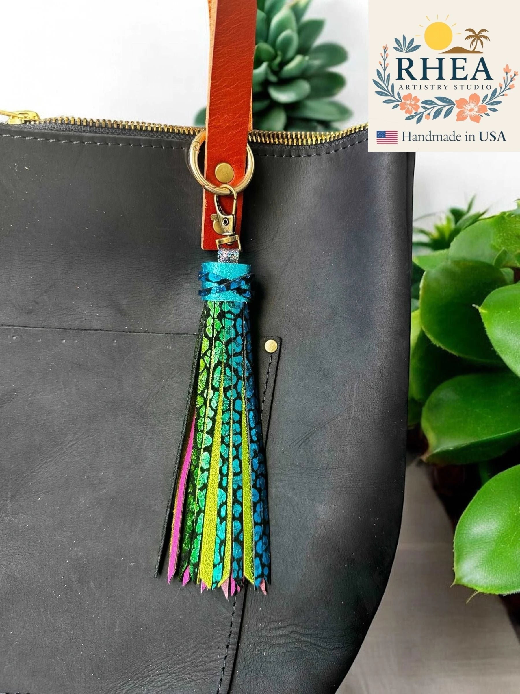 Rhea Artistry Studio 7-inch rainbow chameleon leather tassel keychain with lobster clasp attached to a black leather bag.