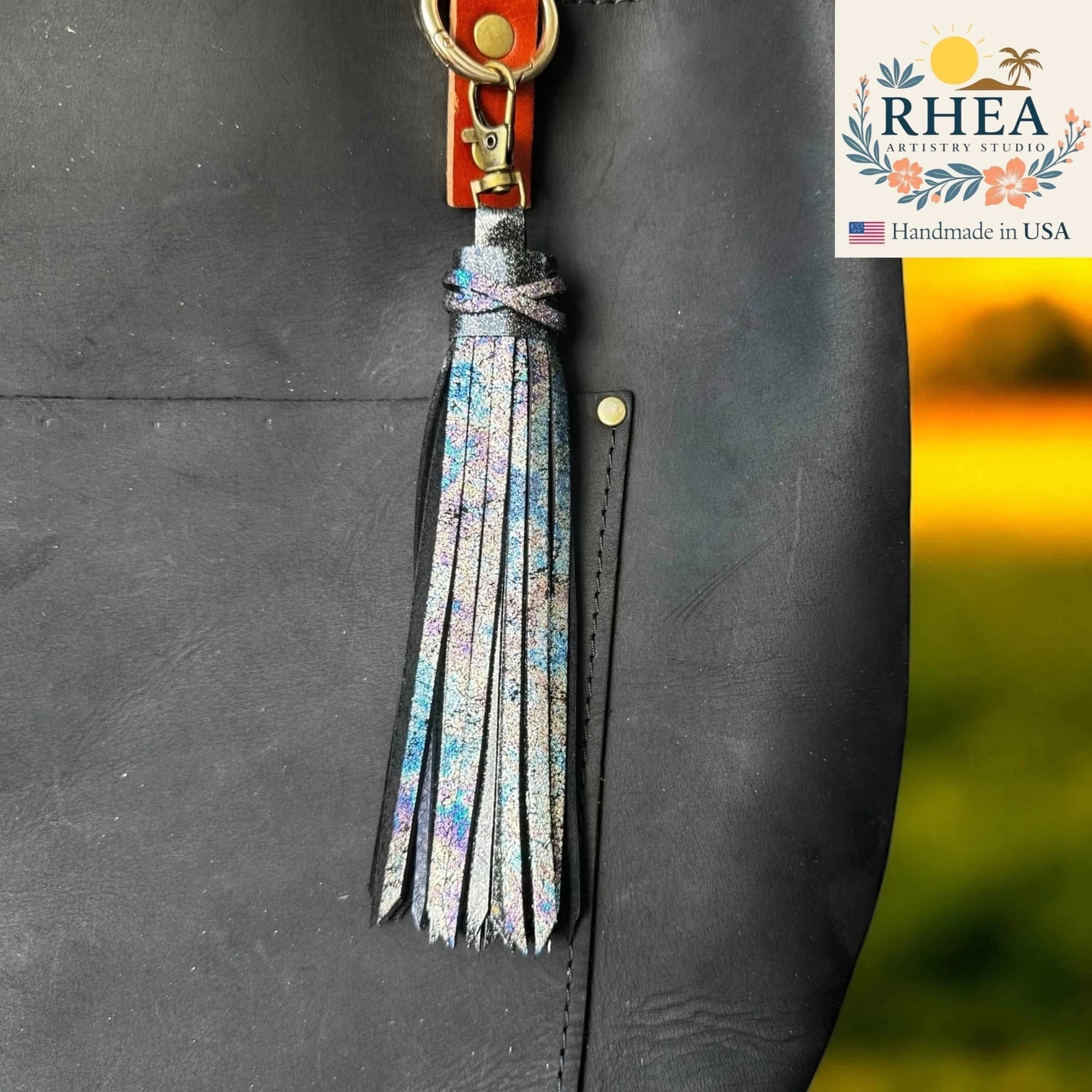 Rhea Artistry Studio handmade 7-inch rainbow chameleon leather tassel keychain with lobster clasp attached to a black tote bag.