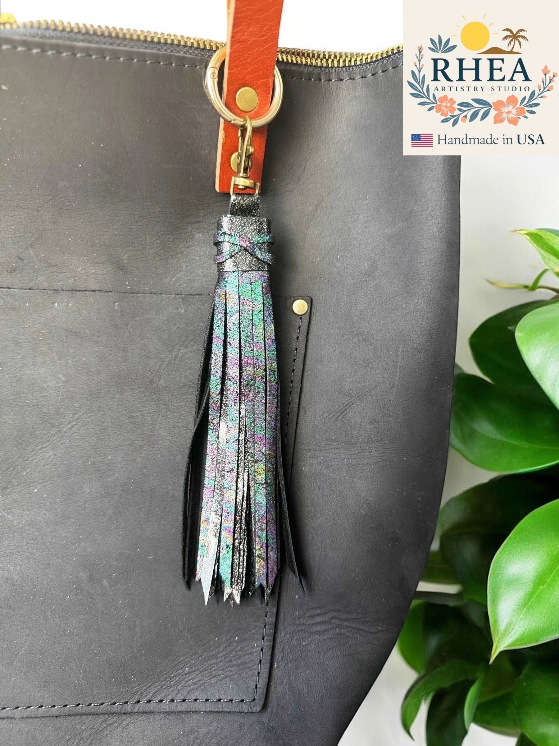 Rhea Artistry Studio 7-inch rainbow chameleon leather tassel keychain with lobster clasp attached to a black leather tote bag.