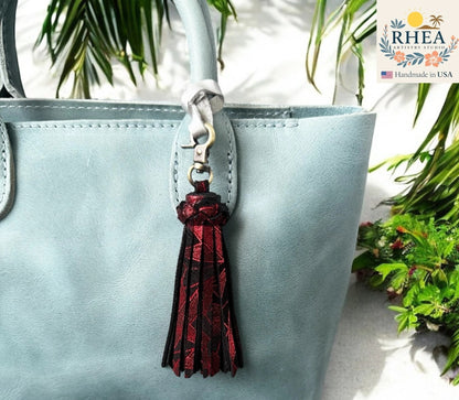 Rhea Artistry Studio handmade red and black leather tassel bag charm with lobster clasp, attached to a light blue tote bag.