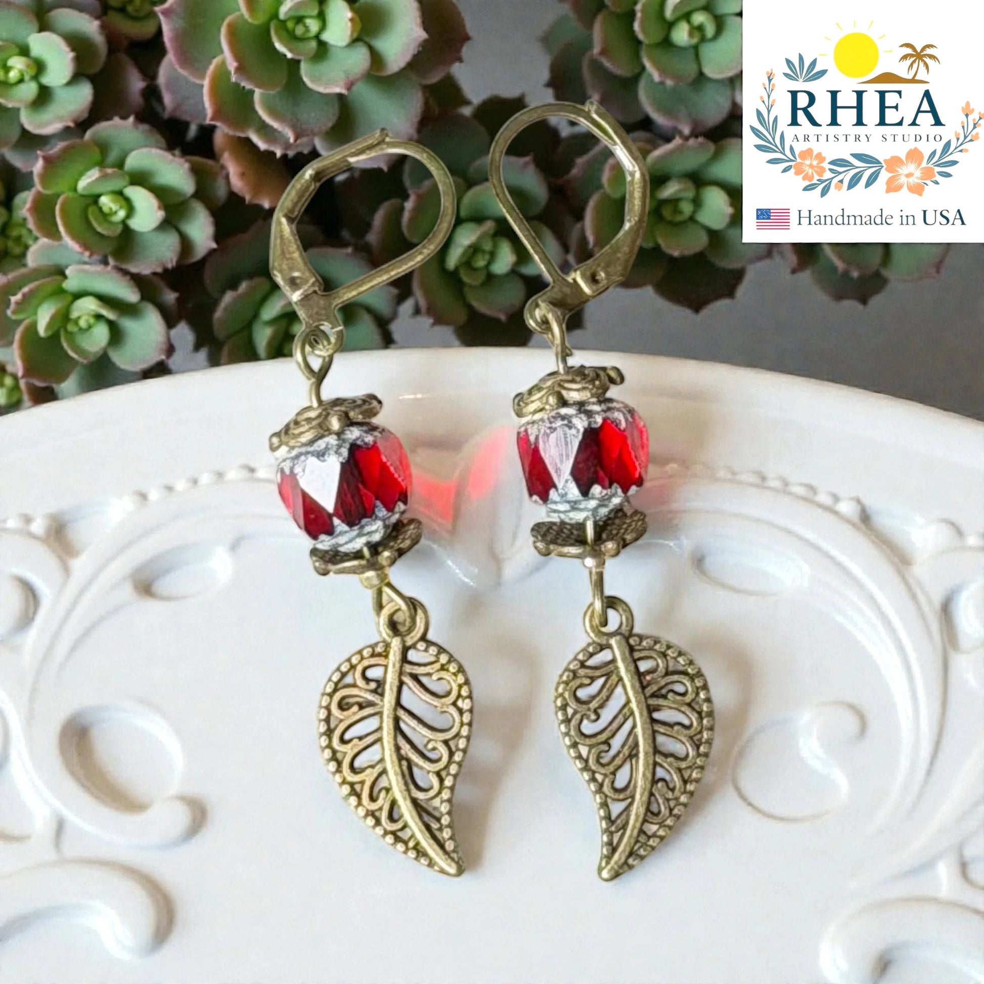 Rhea Artistry Studio handcrafted antique brass leaf drop earrings with faceted red glass beads and Bali-style spacers, 2 inches long.