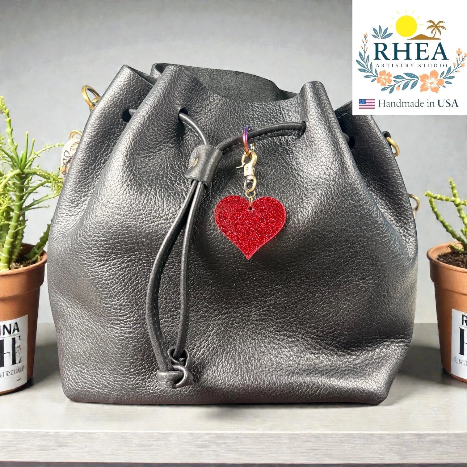 Rhea Artistry Studio red glitter heart charm with antique brass clasp on a black leather bucket bag.