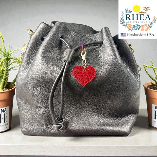 Rhea Artistry Studio red glitter heart charm with antique brass clasp on a black leather bucket bag.