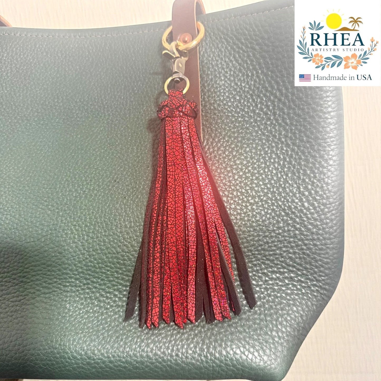 Rhea Artistry Studio handmade red glitter leather tassel keychain with lobster clasp attached to a green pebbled leather bag.