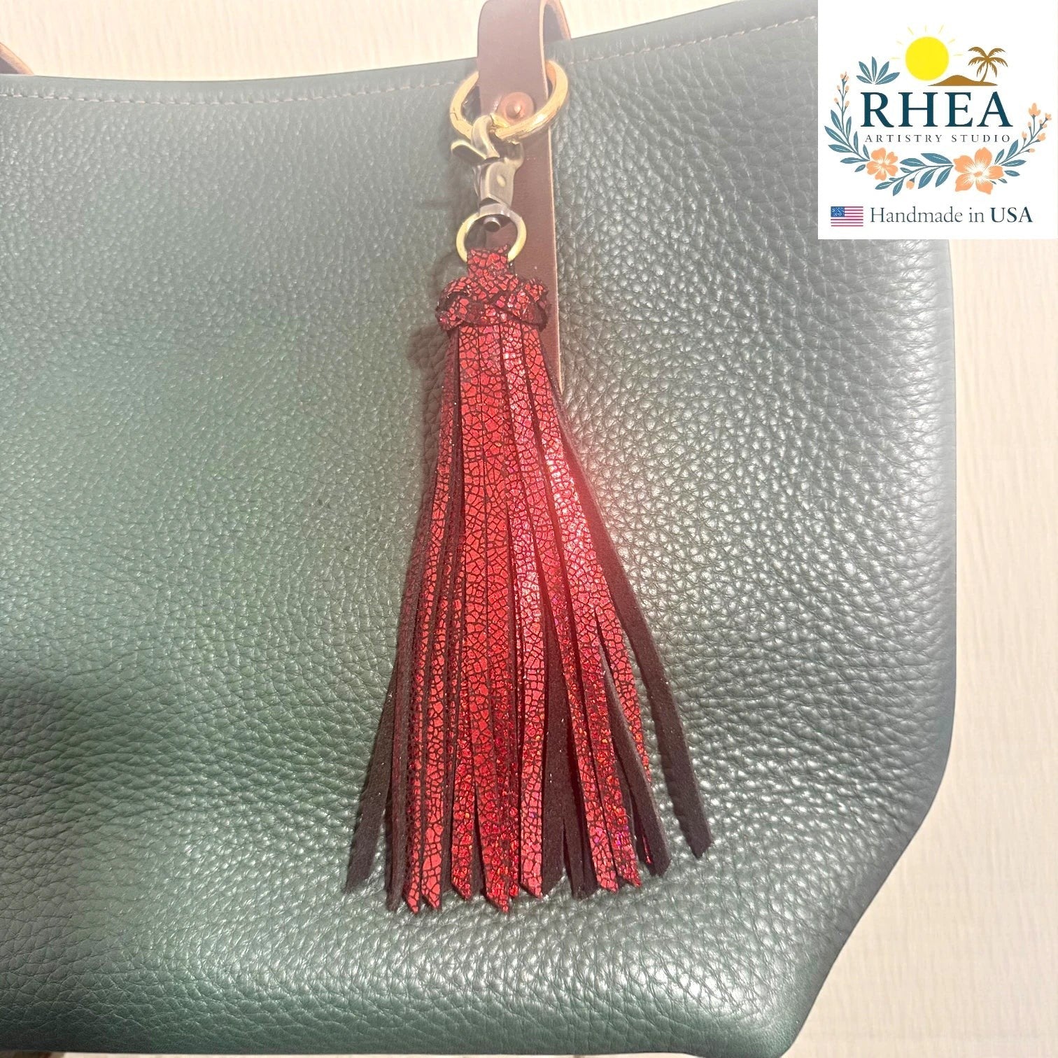 Rhea Artistry Studio handmade red glitter leather tassel keychain with lobster clasp attached to a green pebbled leather bag.