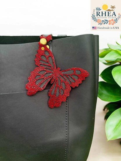Rhea Artistry Studio red leather butterfly charm with metallic finish and gold snap closure attached to a black tote bag.
