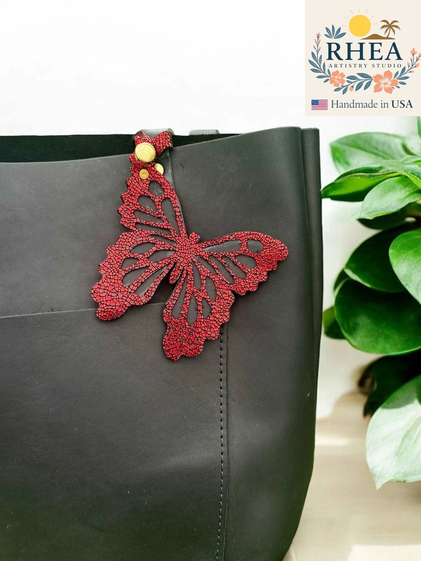 Rhea Artistry Studio red leather butterfly charm with metallic iridescent finish attached to a black tote bag.