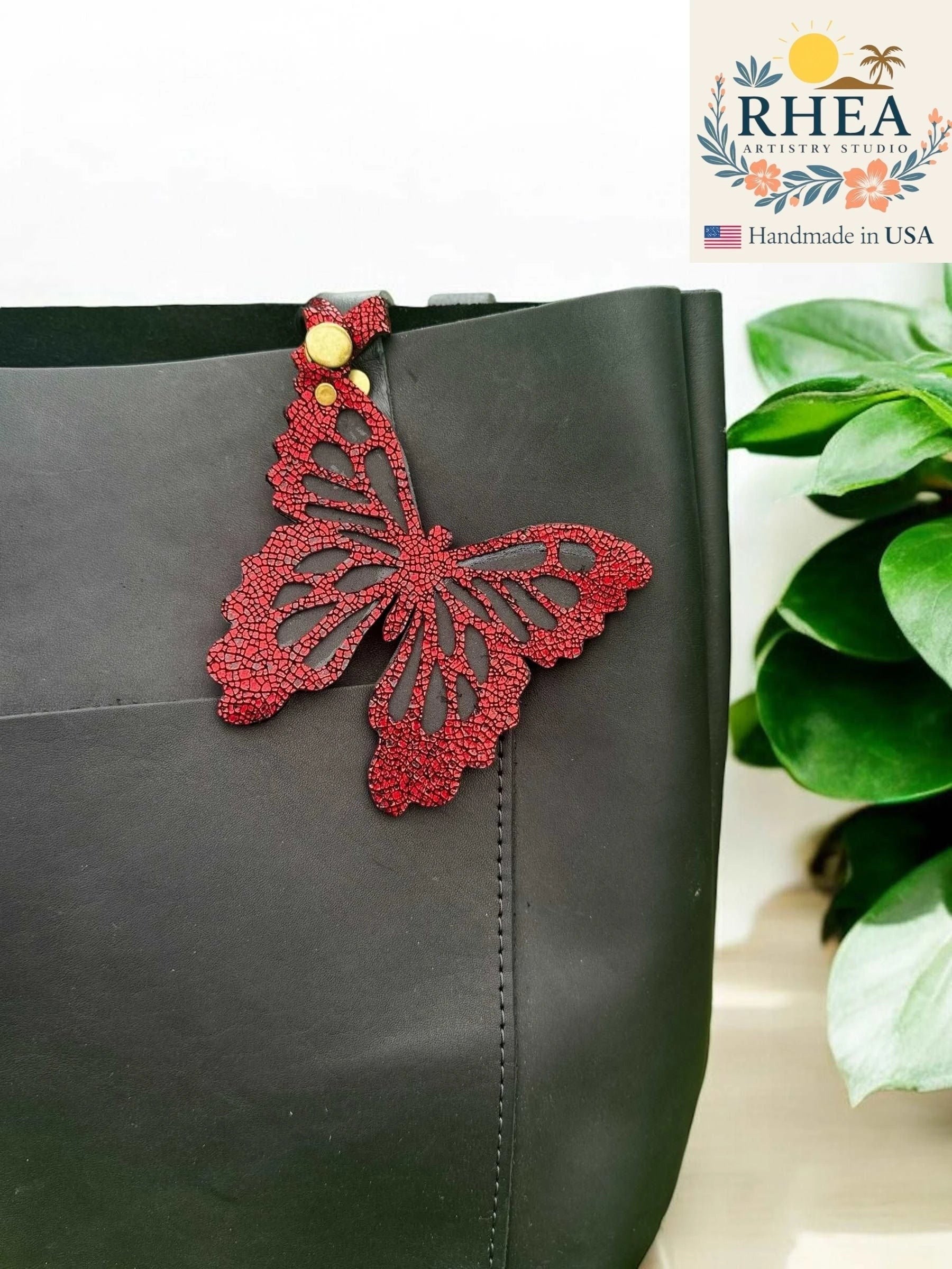 Rhea Artistry Studio red leather butterfly charm with metallic iridescent finish attached to a black tote bag.
