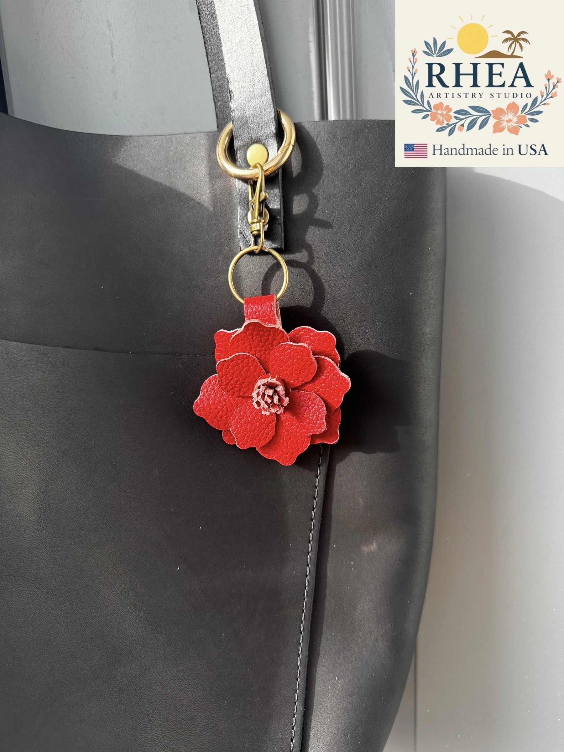 Rhea Artistry Studio red leather flower charm with detailed stamen, attached to a black tote bag with a gold clasp.