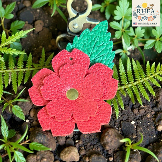 Rhea Artistry Studio red layered leather rose charm with green engraved leaf and brass hardware, set against natural greenery and soil.