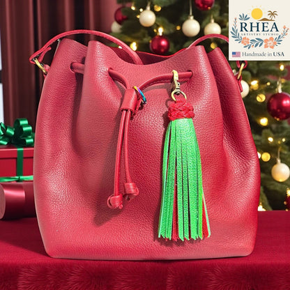 Rhea Artistry Studio red leather bucket bag with a green and red leather tassel charm, antique brass clasp, festive holiday background.