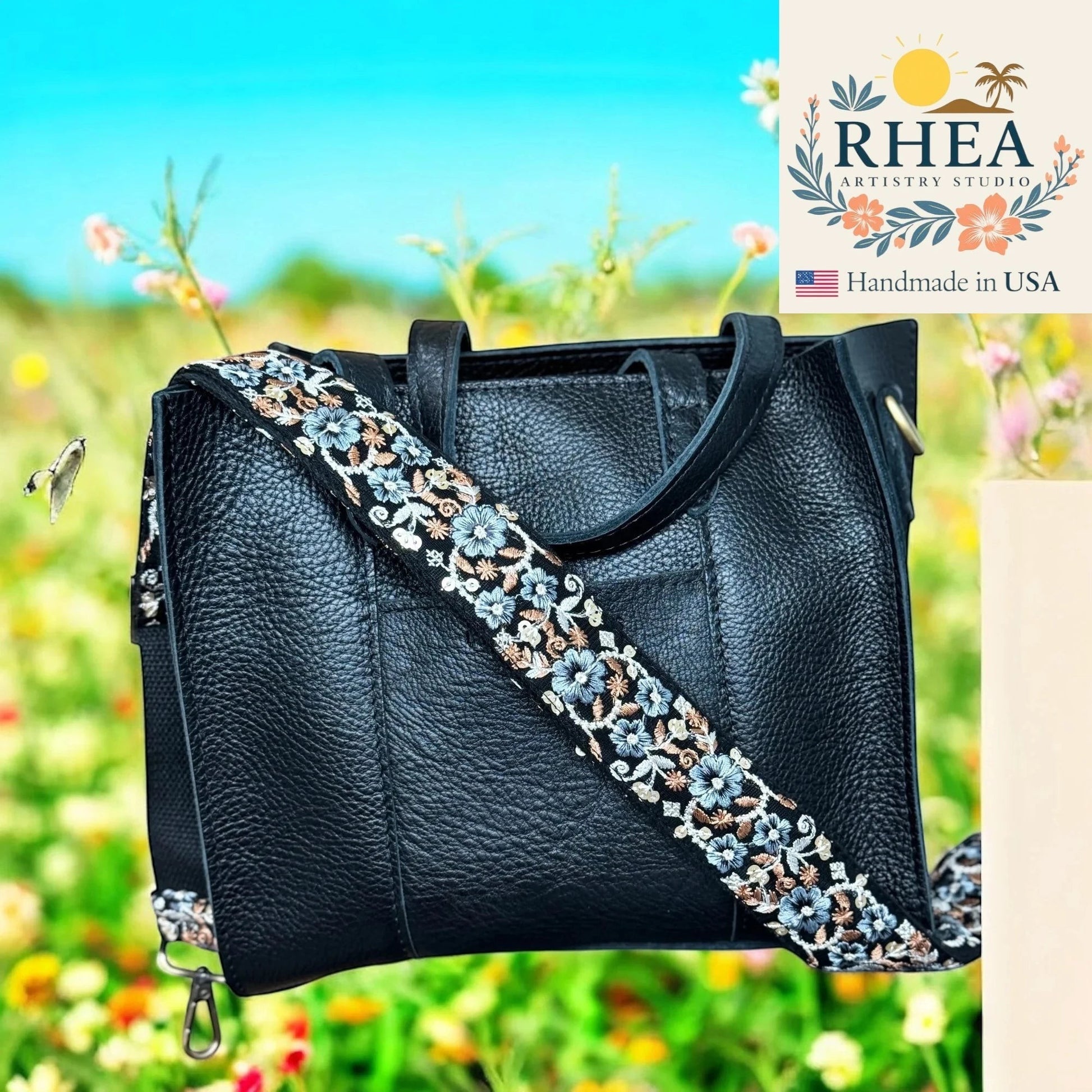 Rhea Artistry Studio black leather handbag with a 1.5-inch seafoam embroidered floral crossbody strap, adjustable length, brushed brass hardware.