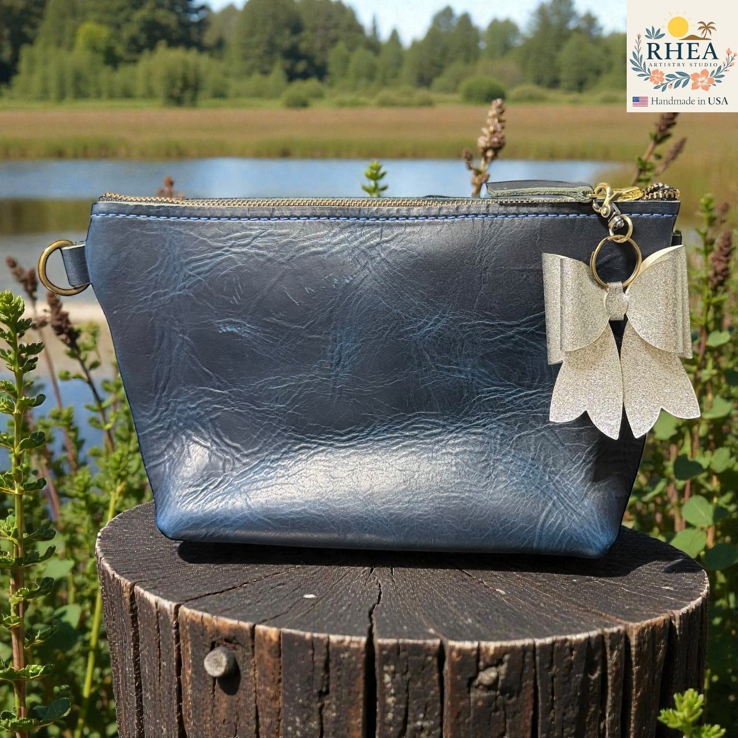 Rhea Artistry Studio silver glitter double bow charm with brass hardware attached to a navy blue textured cosmetic bag.