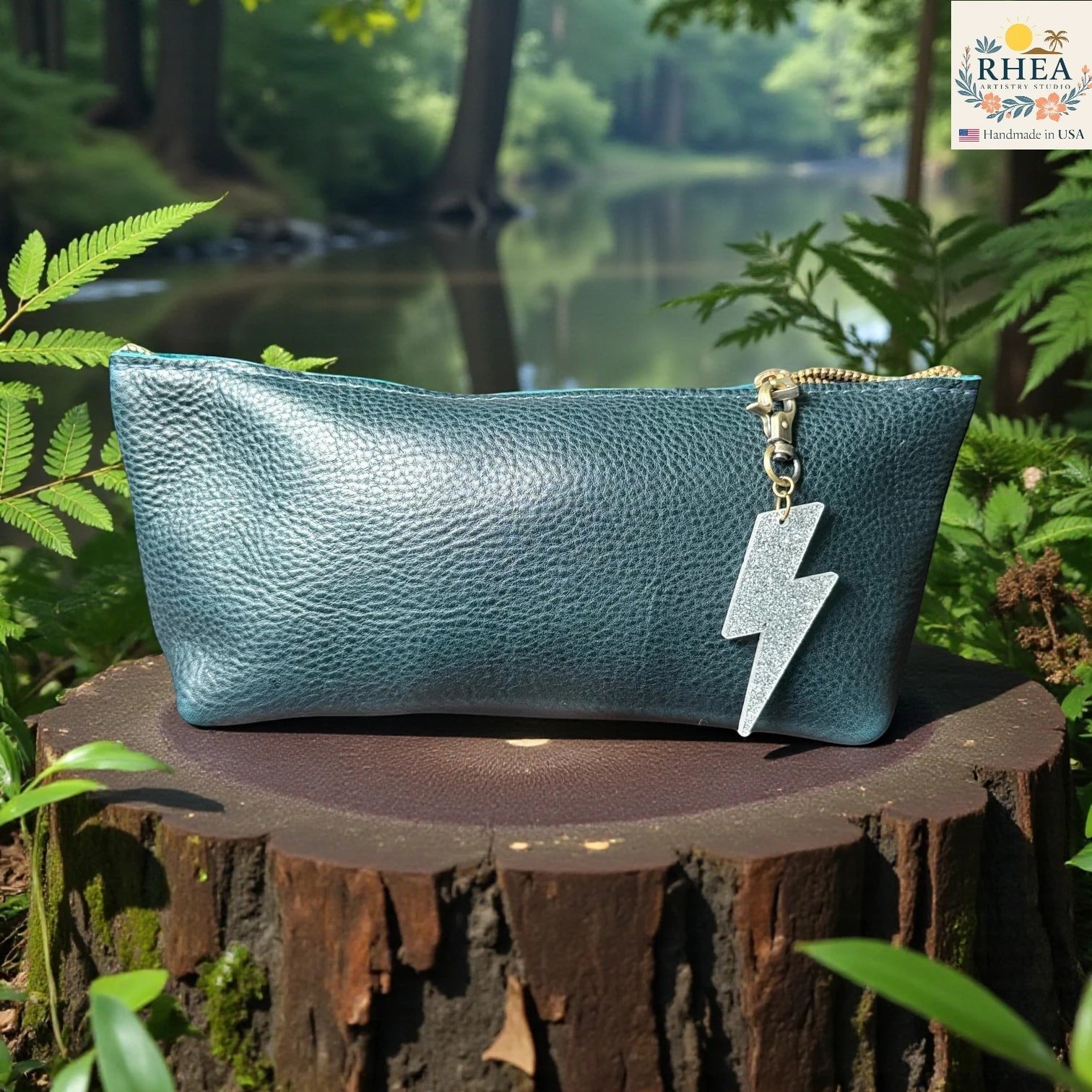 Rhea Artistry Studio silver glitter lightning bolt charm attached to a teal pebbled leather pouch, displayed on a tree stump outdoors.