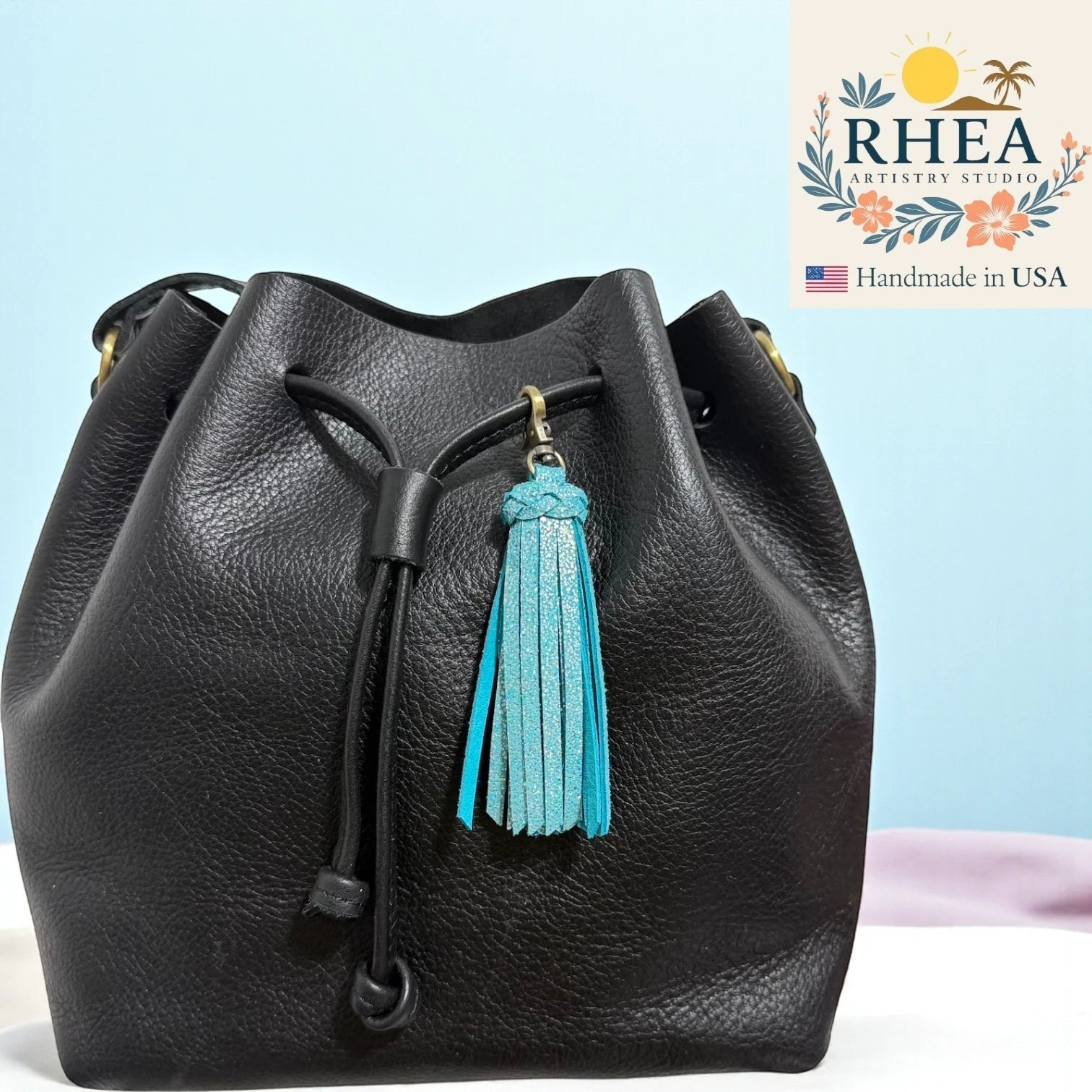 Rhea Artistry Studio handmade sparkly turquoise leather tassel bag charm with antique brass lobster clasp attached to a black leather drawstring bag.