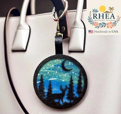 Rhea Artistry Studio hand-painted leather bag charm featuring a starry night sky with a crescent moon, pine trees, and a deer silhouette on vegetable-tanned leather.