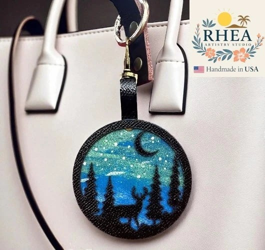 Rhea Artistry Studio hand-painted leather bag charm featuring a starry night sky with a crescent moon, pine trees, and a deer silhouette on vegetable-tanned leather.