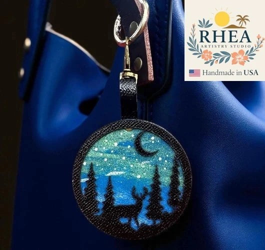 Rhea Artistry Studio hand-painted leather bag charm featuring a starry night sky, crescent moon, pine trees, and a deer silhouette on vegetable-tanned leather.
