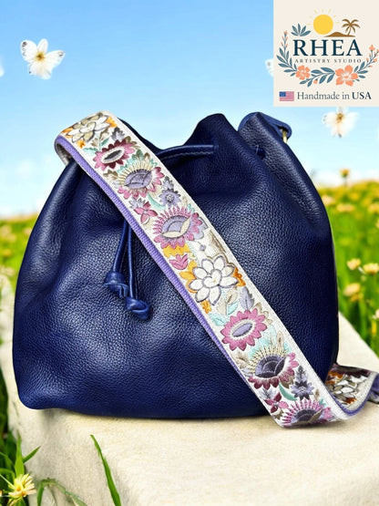 Rhea Artistry Studio navy blue leather bucket bag with a 2-inch wide adjustable crossbody strap featuring colorful floral embroidery and brushed brass hardware.