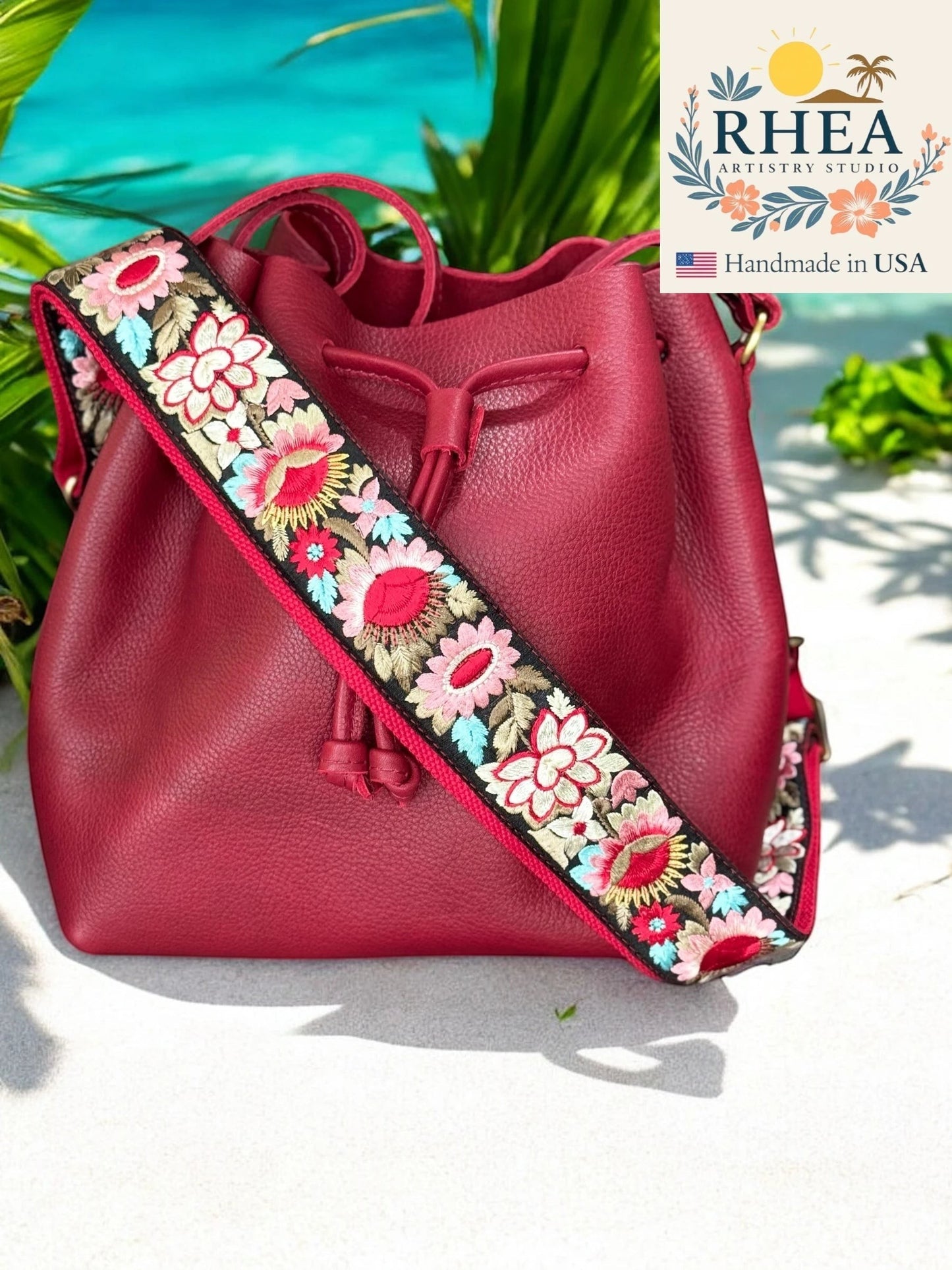 Rhea Artistry Studio red leather bucket bag with a 2-inch wide adjustable crossbody strap featuring vibrant floral embroidery in pink, red, and blue.