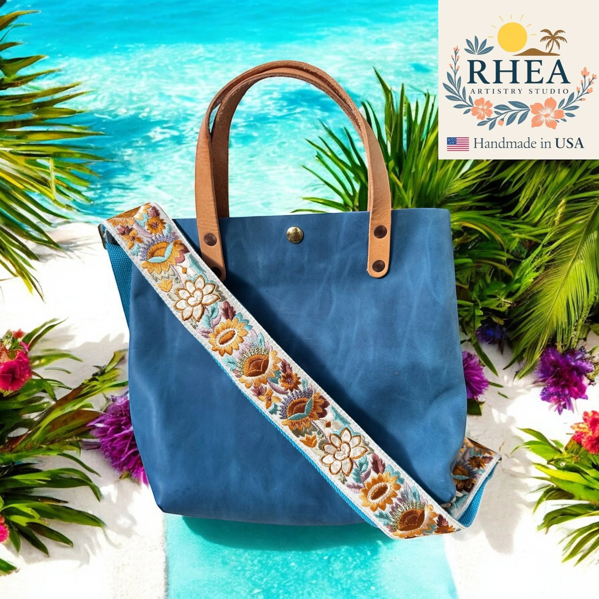 Rhea Artistry Studio blue tote bag with a 2-inch wide adjustable sunflower embroidered strap and leather handles, set against a beach backdrop.