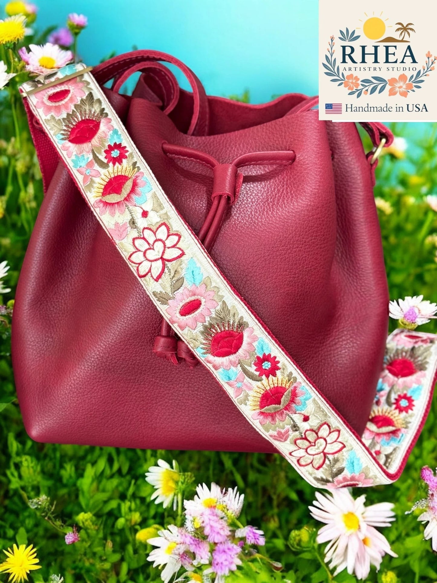 Rhea Artistry Studio 2-inch wide crossbody strap with vibrant floral embroidery in red, pink, and blue on a cream background, attached to a burgundy bucket bag.