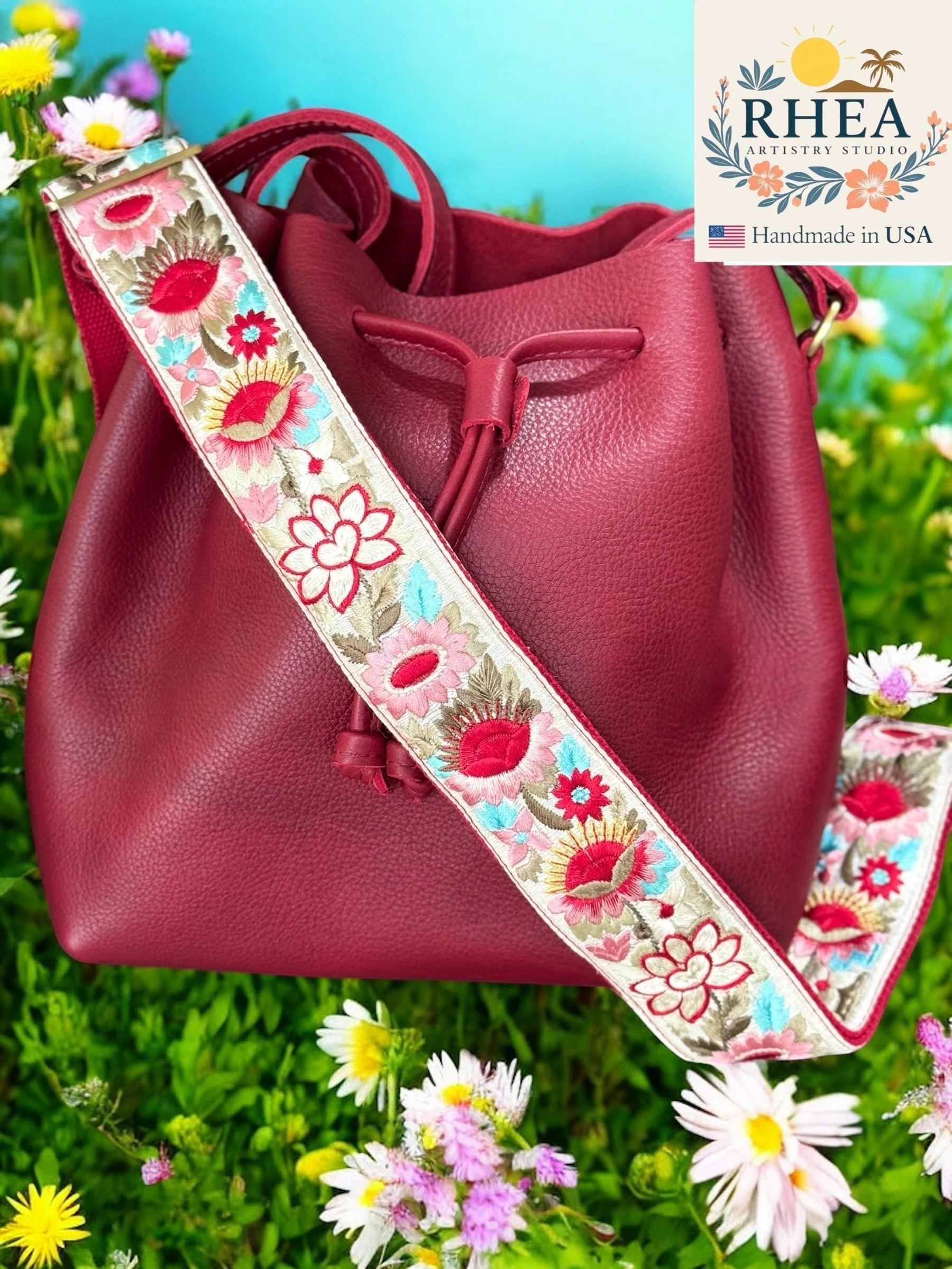 Rhea Artistry Studio 2-inch wide crossbody strap with vibrant floral embroidery in red, pink, and blue on a cream background, attached to a burgundy bucket bag.