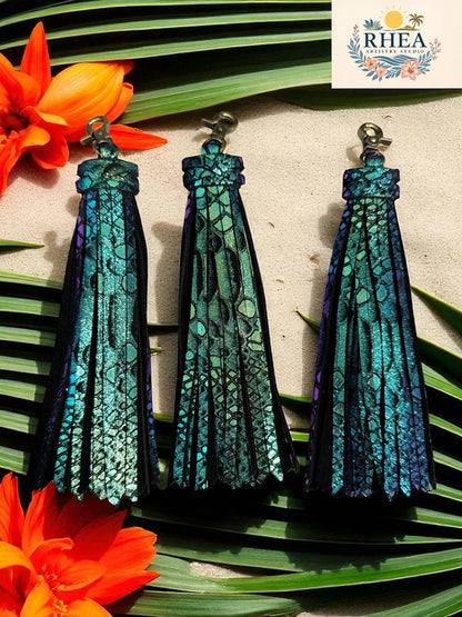 Rhea Artistry Studio handmade teal and purple iridescent leather tassel keychain with lobster clasp, set against a tropical beach background with palm leaves and orange flowers.