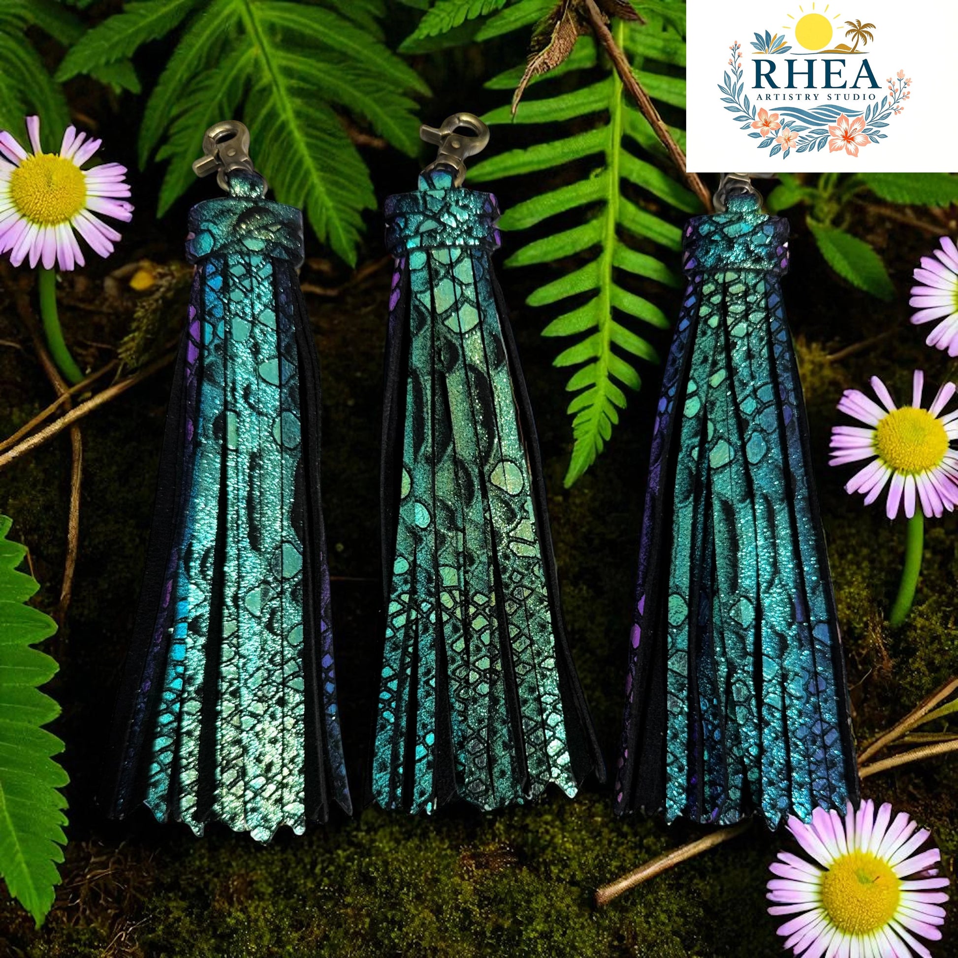 Rhea Artistry Studio teal and purple iridescent leather tassel keychain with lobster clasp, set against a natural backdrop of ferns and flowers.