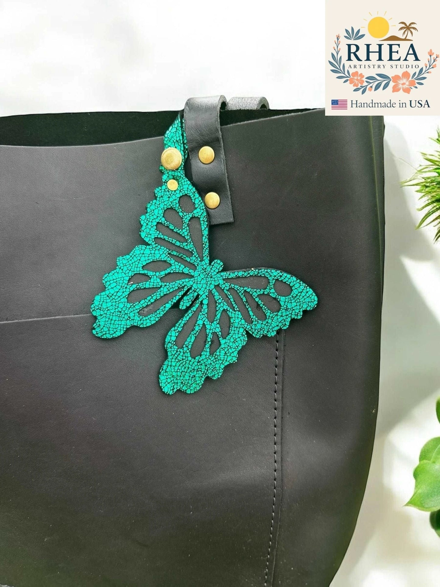 Rhea Artistry Studio teal leather butterfly charm with metallic iridescent finish and gold hardware, attached to a black tote bag.