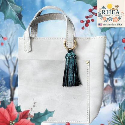 Rhea Artistry Studio handmade teal leather tassel bag charm with gold lobster clasp attached to a white tote bag.