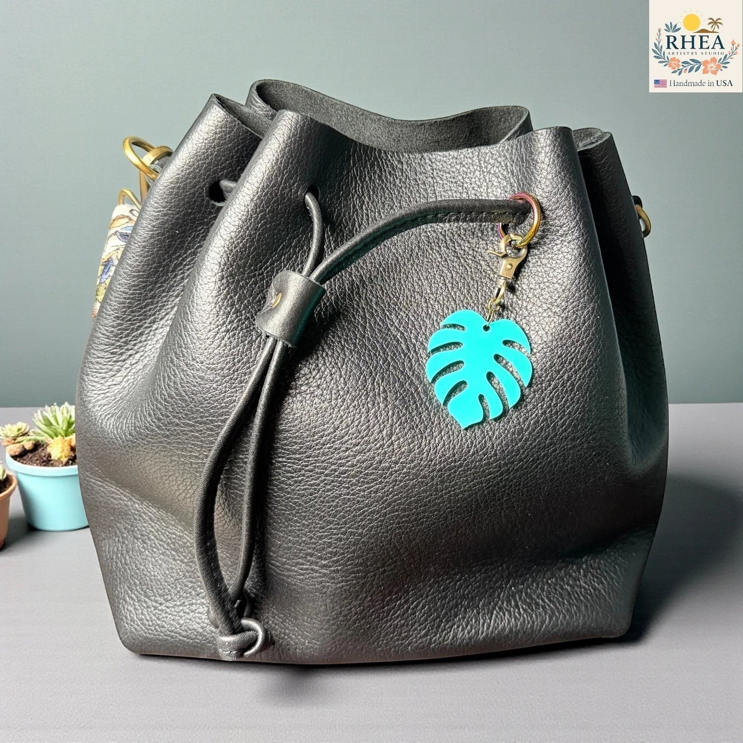 Rhea Artistry Studio teal monstera leaf acrylic charm with antique brass clasp on a black leather drawstring bag.
