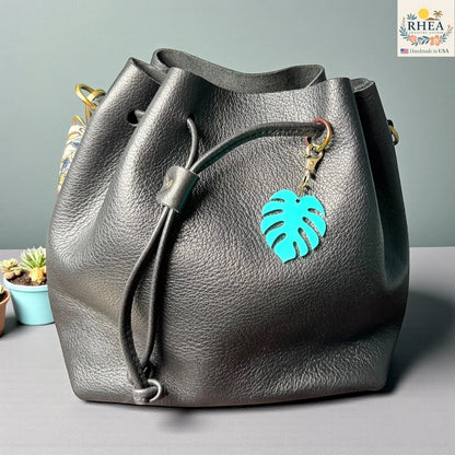 Rhea Artistry Studio teal monstera leaf acrylic charm with antique brass clasp on a black leather drawstring bag.