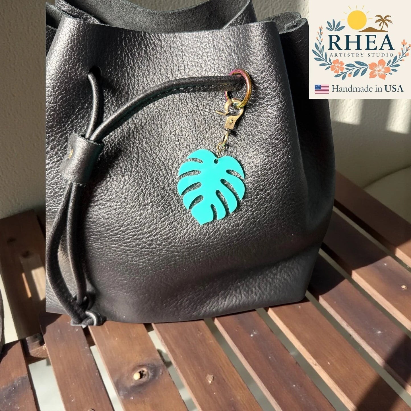 Rhea Artistry Studio teal monstera leaf acrylic charm with antique brass clasp attached to a black leather bag zipper.