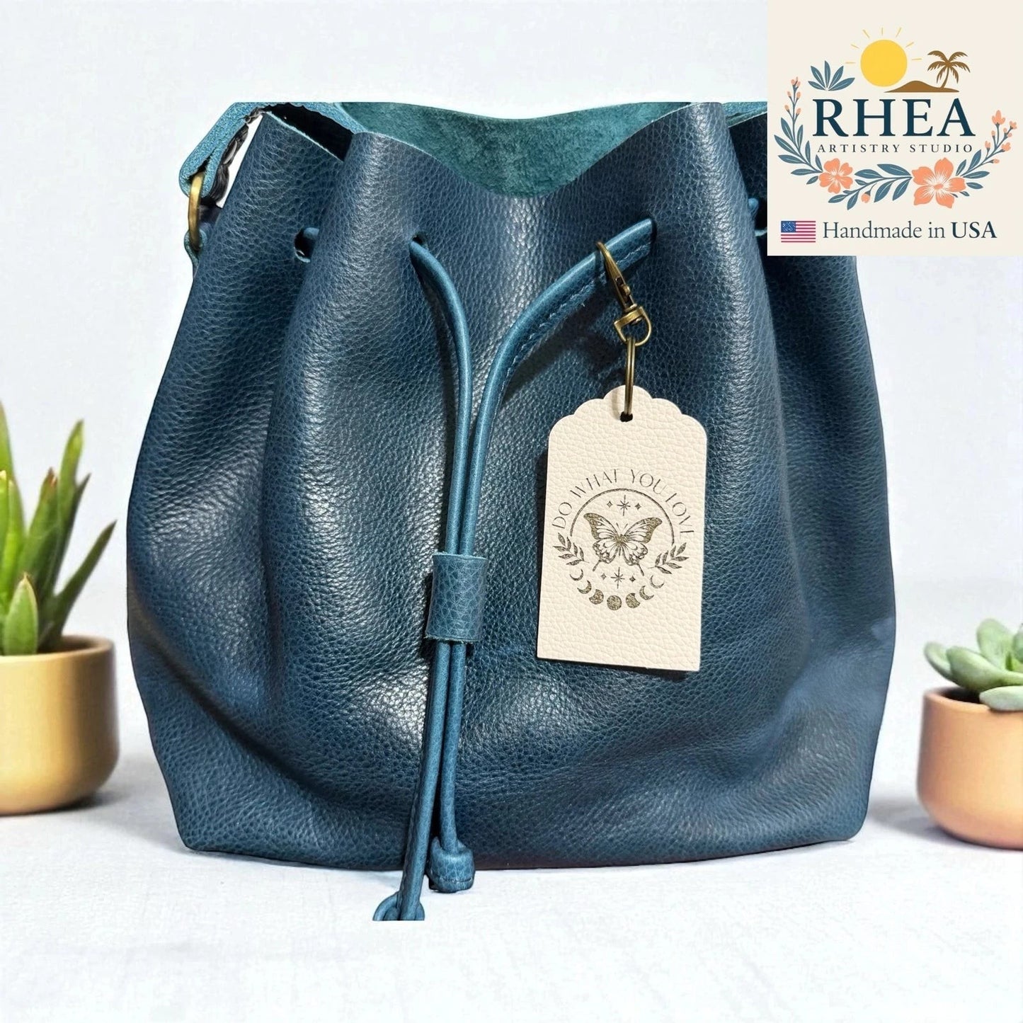 Rhea Artistry Studio teal Saffiano leather bucket bag with a cream leather tag engraved with a butterfly and moon phases.