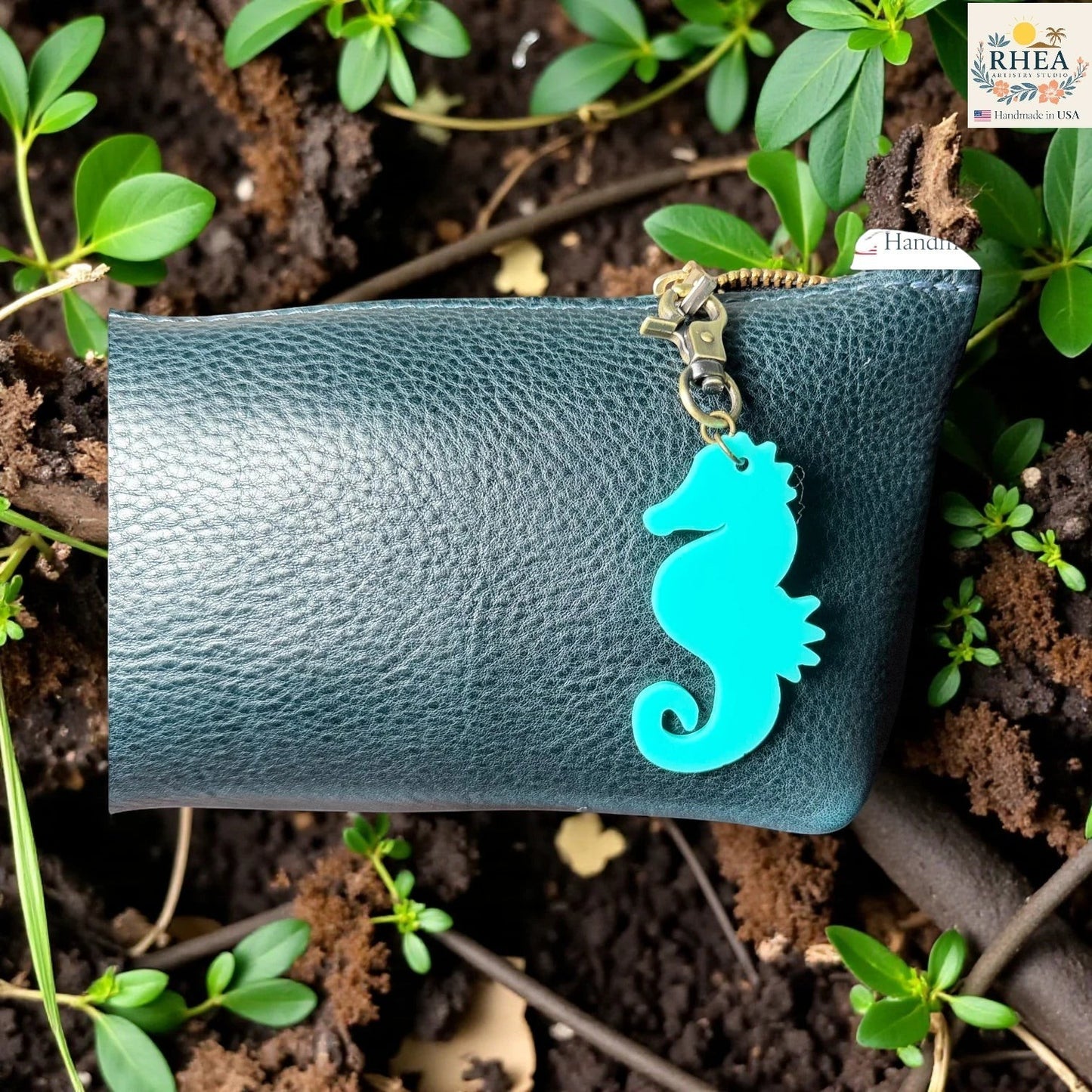 Rhea Artistry Studio teal seahorse acrylic charm with brass clasp on a dark teal leather pouch.