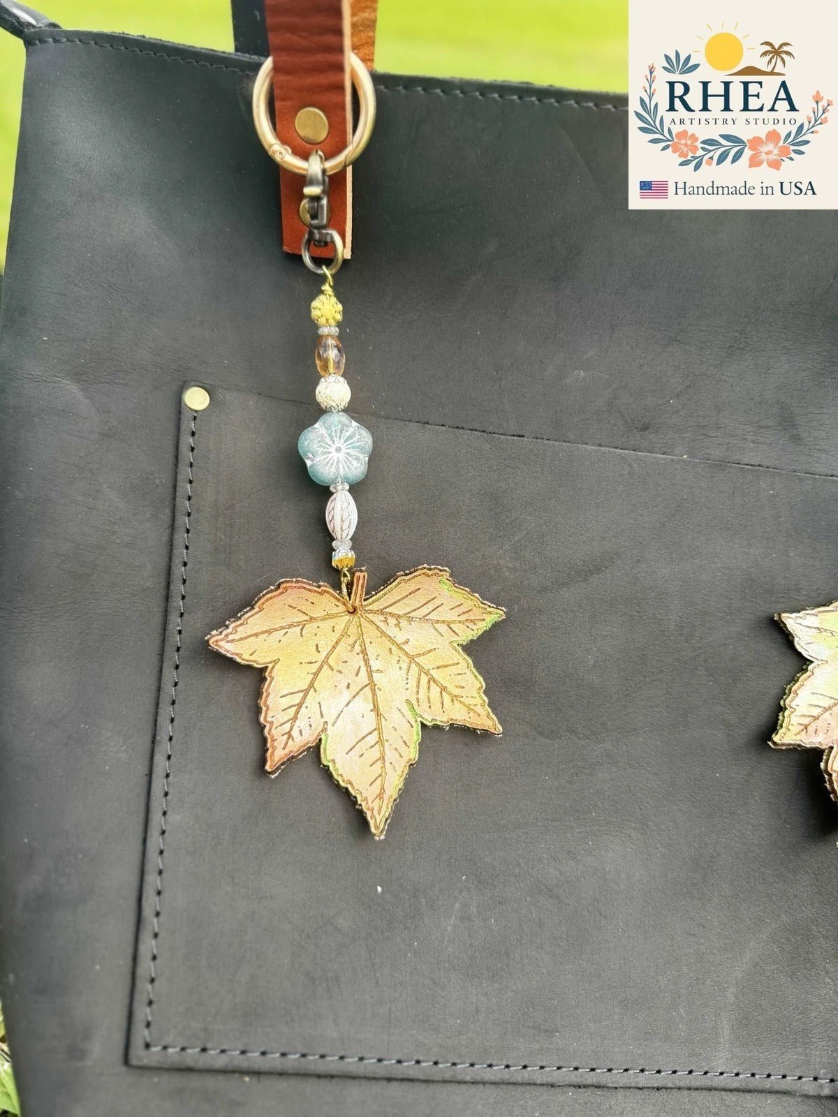 Rhea Artistry Studio teal wave blossom leather charm with brass hardware and Czech glass beads, attached to a black leather tote bag.