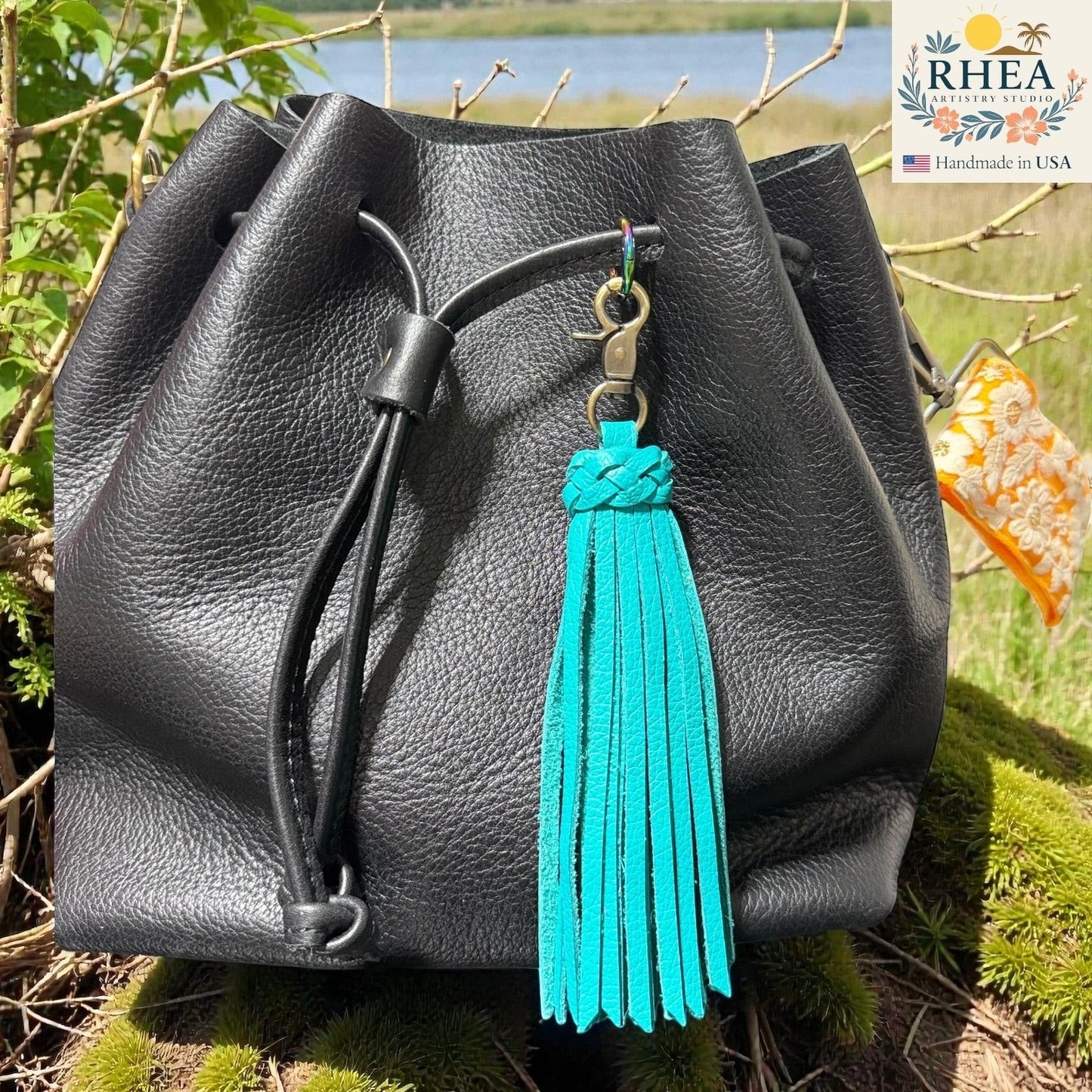 Rhea Artistry Studio turquoise leather tassel bag charm with braided detail and brass clasp, attached to a black leather drawstring bag.