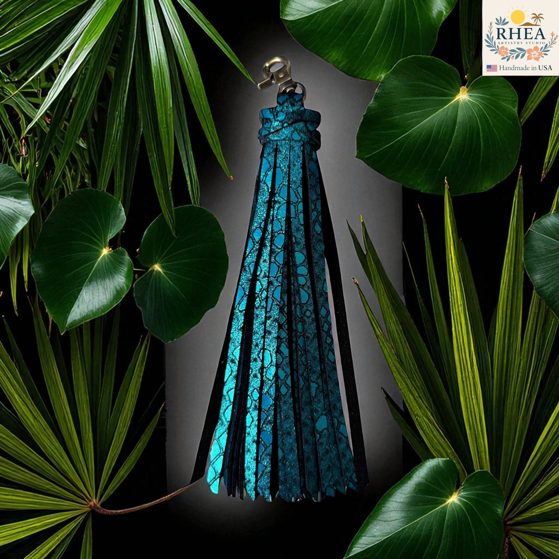 Rhea Artistry Studio turquoise leather tassel keychain with glitter finish and lobster clasp, surrounded by lush green foliage.