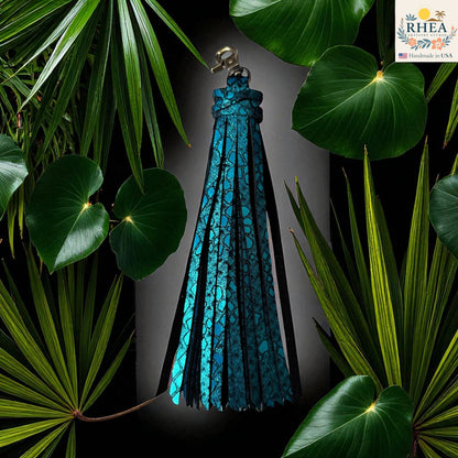 Rhea Artistry Studio turquoise leather tassel keychain with glitter finish and lobster clasp, surrounded by lush green foliage.