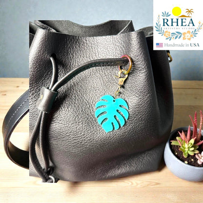 Rhea Artistry Studio turquoise monstera leaf acrylic charm with antique brass clasp on a black leather bag.
