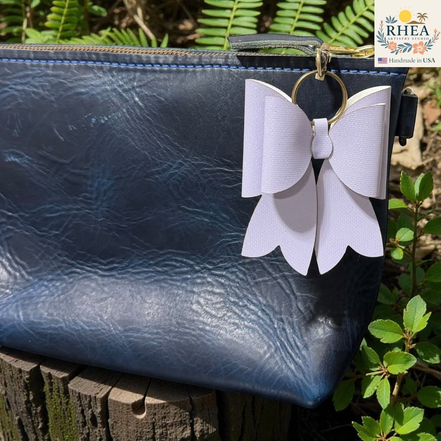 Rhea Artistry Studio white double-layer French bow charm with holographic finish and brass hardware, attached to a navy blue textured leather bag.