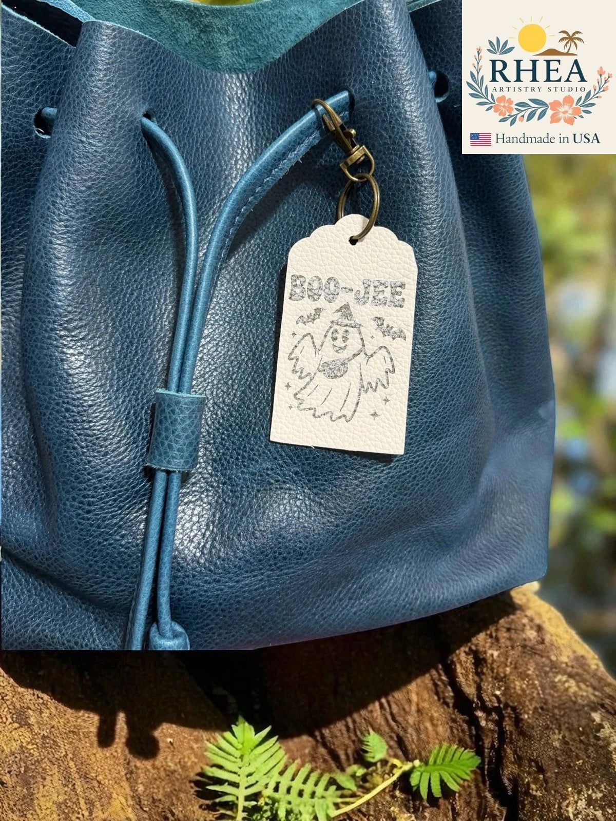 Rhea Artistry Studio white Saffiano leather tag with a ghost graphic and "BOO-JEE" engraving, attached to a blue leather drawstring bag.