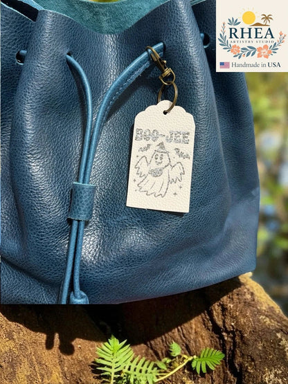 Rhea Artistry Studio white Saffiano leather tag with a ghost graphic and "BOO-JEE" engraving, attached to a blue leather drawstring bag.