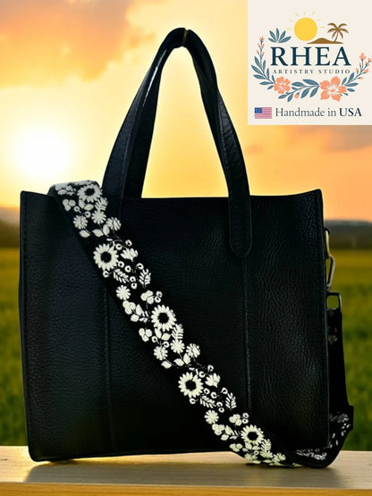 Rhea Artistry Studio black leather tote with a 1.5-inch adjustable crossbody strap featuring white wildflower embroidery.