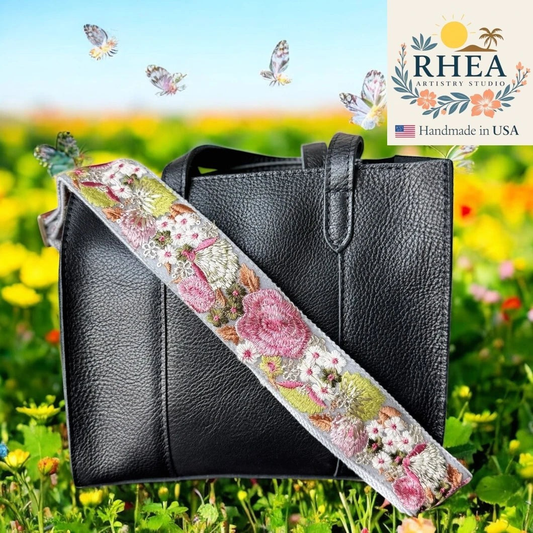 Rhea Artistry Studio 2-inch wide crossbody strap with wildflower embroidery in pink, white, and green on a grey background, attached to a black leather handbag.