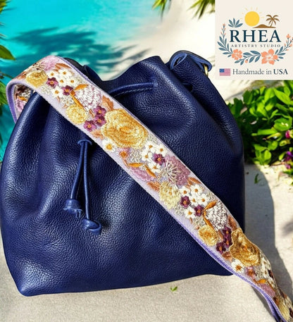 Rhea Artistry Studio navy blue leather bucket bag with a 2-inch wide wildflower embroidered strap in purple, gold, and white.