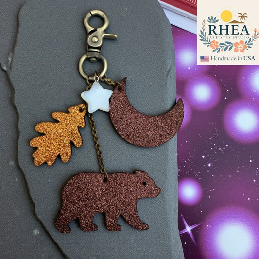 Rhea Artistry Studio woodland charm with glitter leather bear, oak leaf, moon, and opalite star on antique brass hardware.