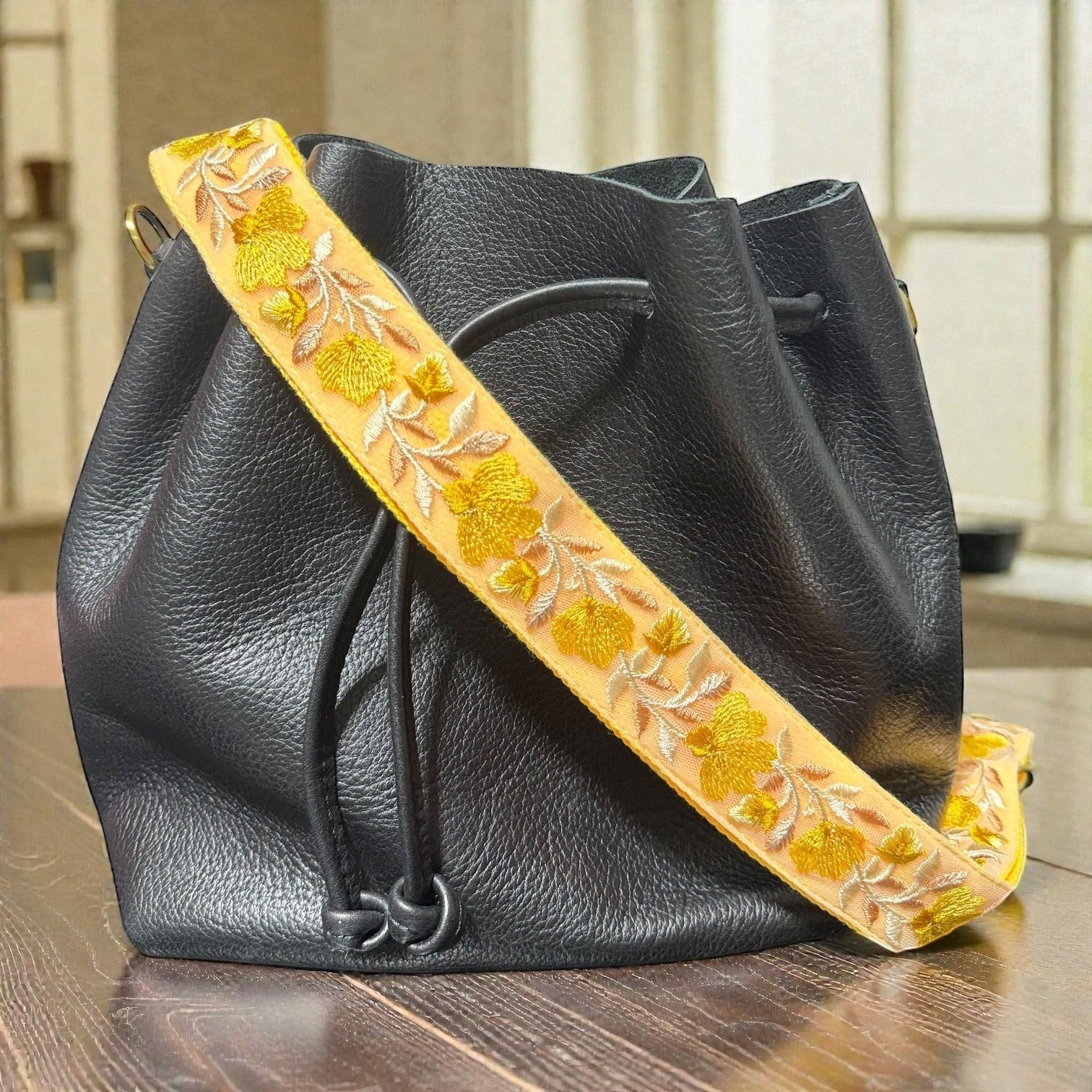 Rhea Artistry Studio yellow embroidered crossbody strap with floral pattern on black leather bucket bag.